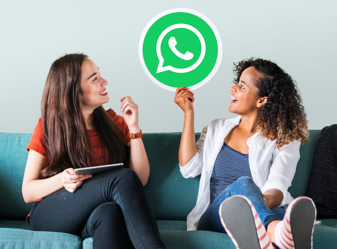 Duas mulheres sentadas em um sofá azul, conversando sobre WhatsApp banido, com uma placa do aplicativo nas mãos.